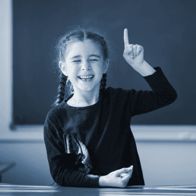Ein Mädchen mit Zöpfen sitzt am Pult vor der Tafel und zeigt mit ihrer linken Hand die Zahl Eins.