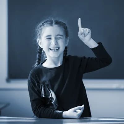 Ein Mädchen mit Zöpfen sitzt am Pult vor der Tafel und zeigt mit ihrer linken Hand die Zahl Eins.